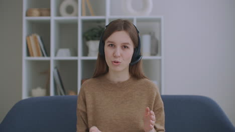 portrait-of-online-consultant-woman-with-headphones-on-head-sitting-at-table-and-talking-to-camera